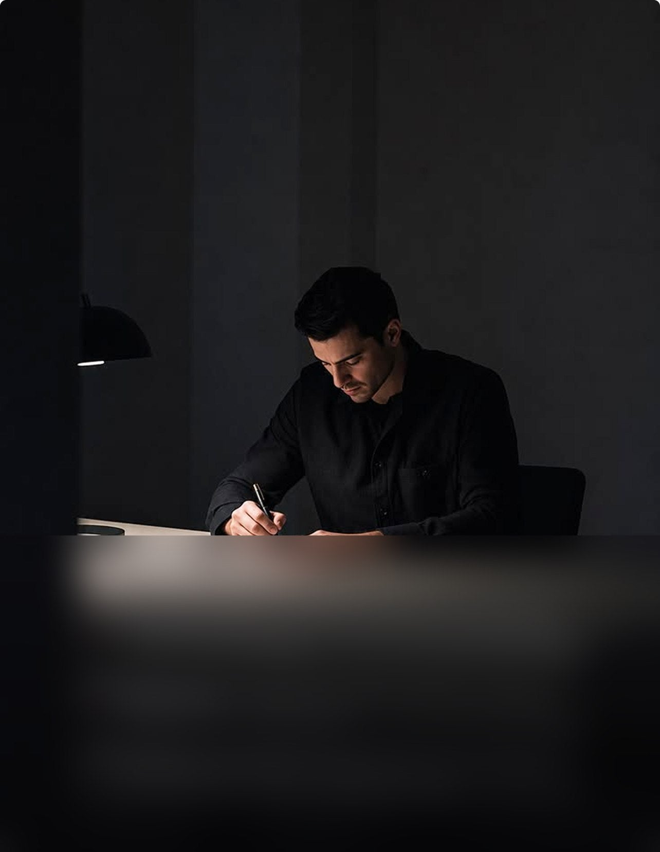 Man writing in a notebook at a desk with a reading lamp at night.